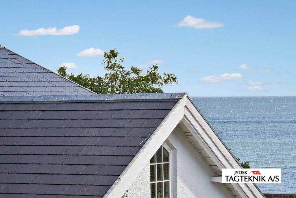 Et mørkt tegltag på et hus, med en hvid kvist og hav i baggrunden under en blå himmel.